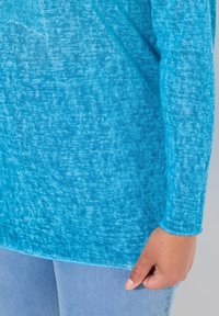 Maglietta a maniche lunghe di colore azzurro chiaro realizzata in un tessuto testurizzato con un sottile effetto ombreggiato; l'orlo è leggermente curvato, con dettagli di cucitura visibili.