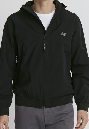 Homme portant une veste à capuche noire zippée sur une chemise blanche et un pantalon gris, debout les bras détendus le long du corps.