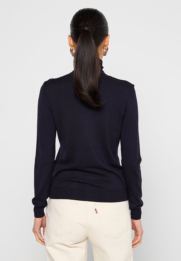 Maglione a coste blu navy con maniche lunghe e una vestibilità aderente, abbinato a pantaloni chiari, caratterizzati da una superficie liscia e un design minimal.