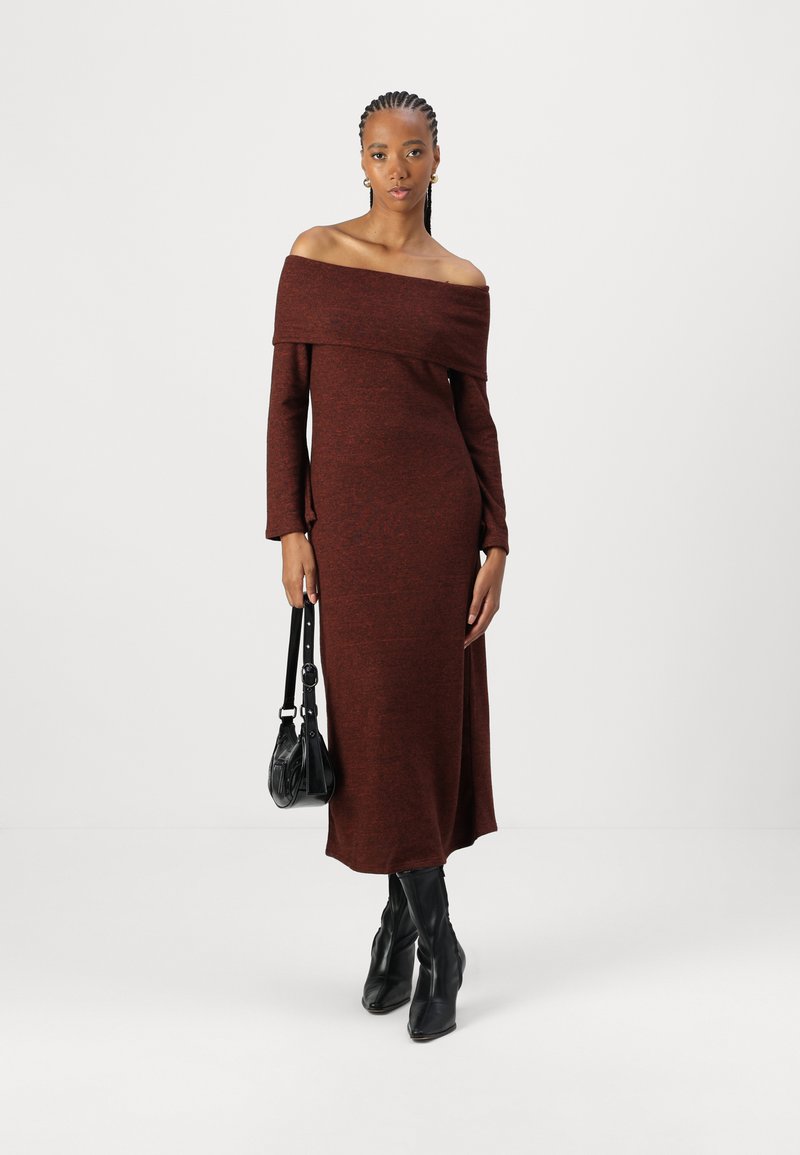 Vestido largo ribeteado en color marrón rojizo, con escote off-the-shoulder y mangas largas, con una hendidura lateral, combinado con botas negras hasta la rodilla y un bolso negro de hombro.