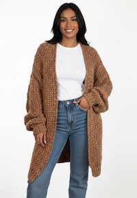 Brązowy dziergany kardigan o luźnym kroju, szerokich rękawach i otwartym przodzie, zestawiony z niebieskimi dżinsami z wysokim stanem oraz białą bluzką.