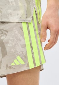 Χέρι δίπλα σε ανοιχτόμπεζ αθλητικό σορτς με νέον κίτρινο λογότυπο και ρίγες Adidas, με διακριτικό αφηρημένο σχέδιο.