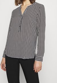 Langærmet mønsterbluse i sort-hvid houndstooth-design, med V-hals og knapdetalje foran. Glat stoftekstur.