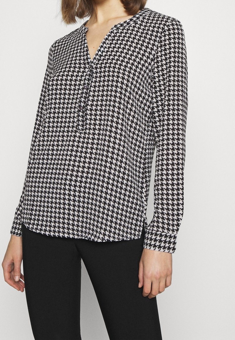 Langærmet mønsterbluse i sort-hvid houndstooth-design, med V-hals og knapdetalje foran. Glat stoftekstur.