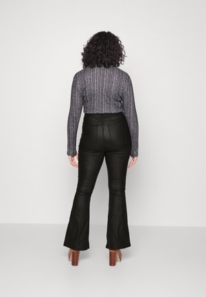 Kobieta o kręconych włosach stoi tyłem, ubrana w szary, prążkowany, długi rękaw, czarne, rozszerzane spodnie z wysokim stanem oraz brązowe buty na obcasie.