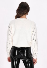 Weißer Strickpullover mit langen Ärmeln, verziert mit silbernen Pailletten an den Ärmeln, kurzes Design, kombiniert mit glänzenden schwarzen Hosen.