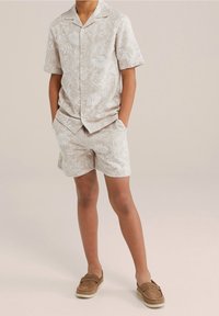 Krátký rukáv, světle béžová košile na knoflíky s bílým tropickým listovým potiskem, spárovaná s odpovídajícími šortkami a hnědými nazouvacími botami.