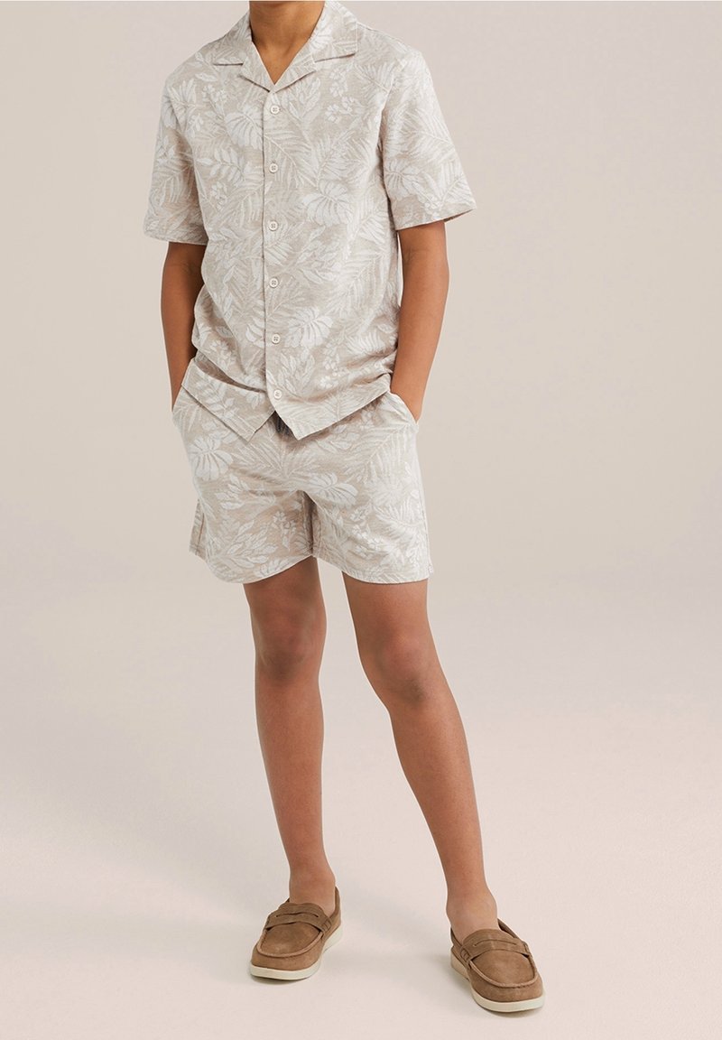 Krátký rukáv, světle béžová košile na knoflíky s bílým tropickým listovým potiskem, spárovaná s odpovídajícími šortkami a hnědými nazouvacími botami.