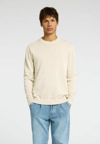 Hellbeige Pullover aus weichem Material mit Rundhalsausschnitt und langen Ärmeln, ausgestattet mit gerippten Bündchen und Saum. Kombiniert mit hellblauen Jeans.