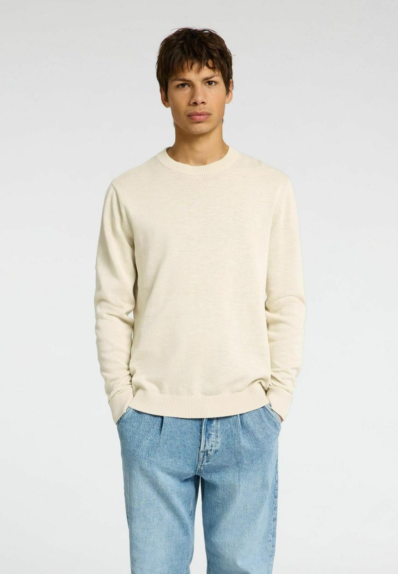Hellbeige Pullover aus weichem Material mit Rundhalsausschnitt und langen Ärmeln, ausgestattet mit gerippten Bündchen und Saum. Kombiniert mit hellblauen Jeans.