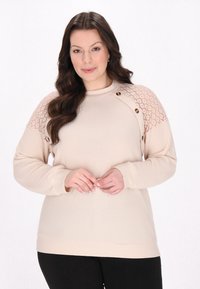 Beige Pullover mit geripptem Textur, Spitze auf den Schultern und zwei dekorativen Knöpfen. Lange Ärmel und Rundhalsausschnitt. Schwarze Hose.