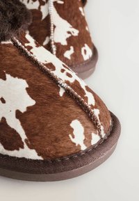 Braun und weiß gefleckte Hausschuhe mit einer weichen Felltextur, kontrastierenden Nähten und einer braunen Gummisohle. Rundliche Zehenform.