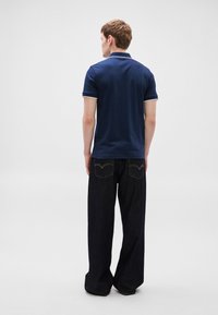 Tricou polo bleumarin cu guler alb și margini la mâneci, asortat cu blugi largi de denim închis la culoare, având buzunare în spate și cusături galbene.