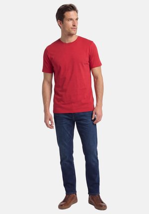 Man staat en draagt een rood T-shirt met korte mouwen, donkerblauwe spijkerbroek en bruine veterschoenen, terwijl hij opzij kijkt.