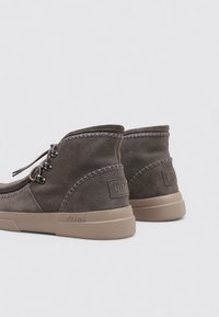 Bottines en suède gris avec une semelle en caoutchouc. Présente des coutures décoratives autour de la cheville et un logo sur le talon. Lacets à l'avant.