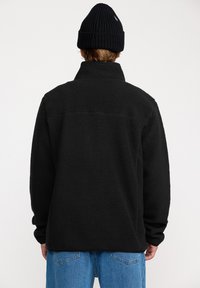 Svart fleecejacka med hög krage, strukturerad yta och lös passform. Bärs med lösa blå jeans och en svart stickad mössa.
