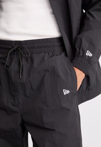 Črne lahke joggers hlače z elastičnim pasom, nastavljivo vezalko in majhnimi belimi logotipnimi detajli ob strani in na robu.