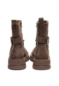 Botas de ante marrón hasta el tobillo con una suela de goma texturizada, que presentan un cierre lateral con cremallera y una correa ajustable con detalle de hebilla metálica.
