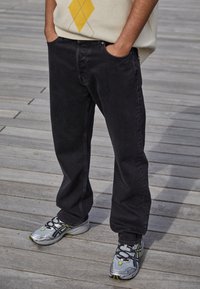 Osoba ubrana w czarne dżinsy, srebrno-żółte sneakersy i jasny sweter w żółty diamentowy wzór, stojąca na drewnianych deskach.