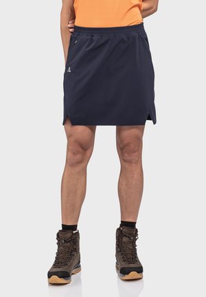 Marineblauer Outdoor-Rock mit elastischem Bund, zwei seitlichen Schlitzen und kleinem Logo, kombiniert mit braunen Wanderschuhen und dunklen Socken.