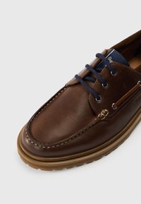 Brązowy skórzany but z zaokrąglonym noskiem, niebieskimi sznurowadłami i beżową gumową podeszwą. Charakteryzuje się widocznym przeszyciem i metalowymi oczkami.