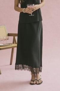 Donna in un top senza maniche nero e gonna lunga con orlo di pizzo tiene un bicchiere a coppa con una bevanda rosa, in piedi vicino a una sedia di legno con un vassoio.