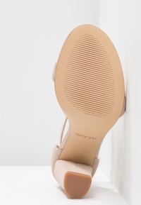 Chaussure mi-hauteur en suède beige avec un talon bloc, une semelle texturée, et la marque "ALDO" au dos, positionnée debout contre un mur blanc.