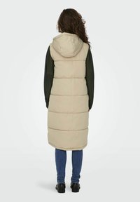 Beige Steppweste mit Kapuze, die ein gestepptes Design aufweist. Getragen über einem schwarzen Oberteil und blauen Jeans, ergänzt durch schwarze Schuhe.