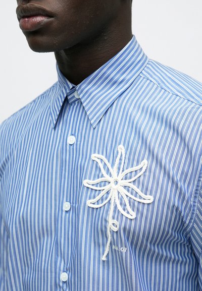 Vīrietis valkā zilbalts svītrains krekls ar pogām un baltu izšūtu ziedu kreisajā krūšu daļā.