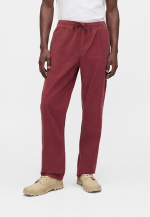 Homme portant un pantalon rouge ample avec une taille élastique et un cordon, des chaussures beige à lacets, et une chemise blanche, debout devant un fond uni.