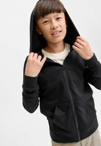Garçon aux cheveux bruns courts portant un sweat à capuche noir zippé sur une chemise claire, tenant la capuche avec les deux mains, pantalon beige visible.