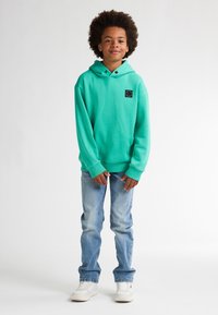 Felpa verde menta con una toppa nera, maniche lunghe e tasca a marsupio, abbinata a jeans azzurri e sneakers bianche.