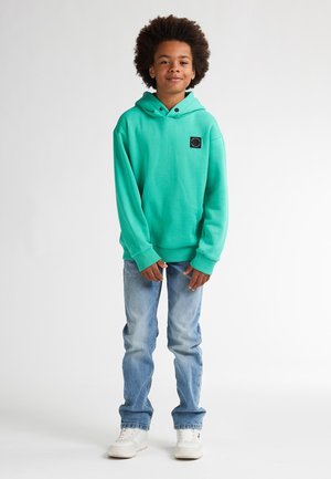 Mintgroene hoodie met een zwart logo-patch, lange mouwen en een kangoeroezak, gecombineerd met lichtblauwe jeans en witte sneakers.