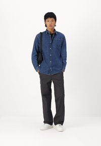 Camisa de mezclilla de botones en azul oscuro, con un único bolsillo en el pecho; combinada con pantalones cargo negros de corte holgado y zapatillas blancas.
