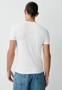 Balta īsroku T-krekls ar apaļu apkakli, pieguloša dizaina un gludas tekstūras, valkāts ar gaiši zilām džinsa biksēm. Nav redzamu logotipu vai rakstu.