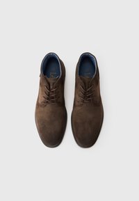 Un par de zapatos para hombre de ante marrón con cordones y forro interior azul, vistos desde arriba sobre un fondo blanco.