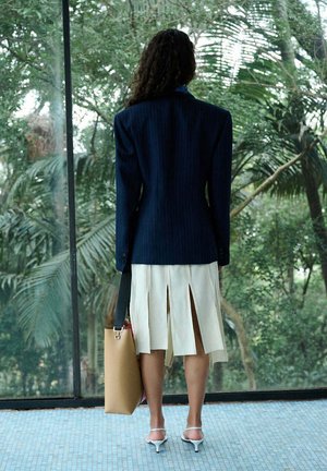 Osoba o kręconych włosach, ubrana w granatową marynarkę, białą plisowaną spódnicę midi, białe szpilki, trzyma beżową torbę na ramię, stoi przed dużym oknem z roślinnością.