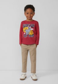 Rotes Langarmshirt mit Grafikdruck und gelben Smiley-Gesichtern, kombiniert mit beigen Hosen und weißen Sneakern mit silbernen Akzenten.