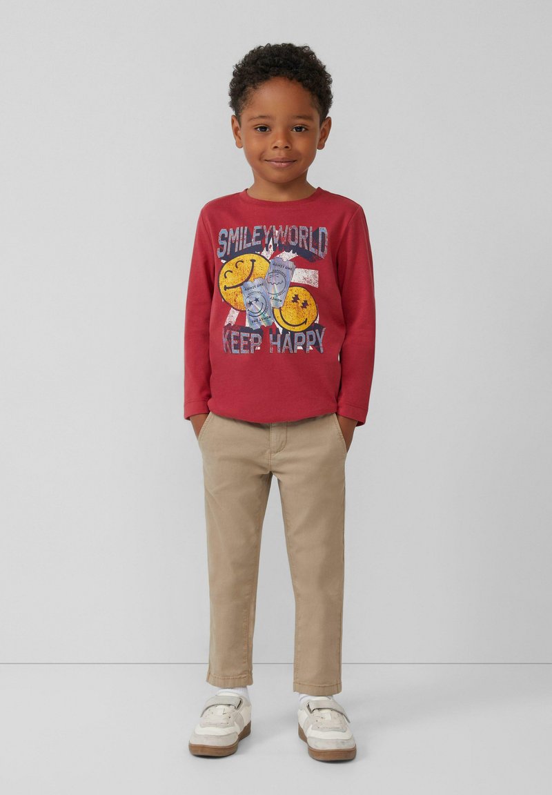 Rotes Langarmshirt mit Grafikdruck und gelben Smiley-Gesichtern, kombiniert mit beigen Hosen und weißen Sneakern mit silbernen Akzenten.