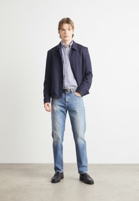Mornarska jakna preko črtaste srajce, svetlo modri jeans s staranim videzom in črne čevlje. Model stoji ob enobarvni ozadju.