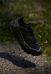Zwarte Salomon trailrunningschoen die boven een rotsachtige ondergrond zweeft, met een achtergrond van groene bladeren. De zoolstructuur en het vetersysteem zijn zichtbaar.