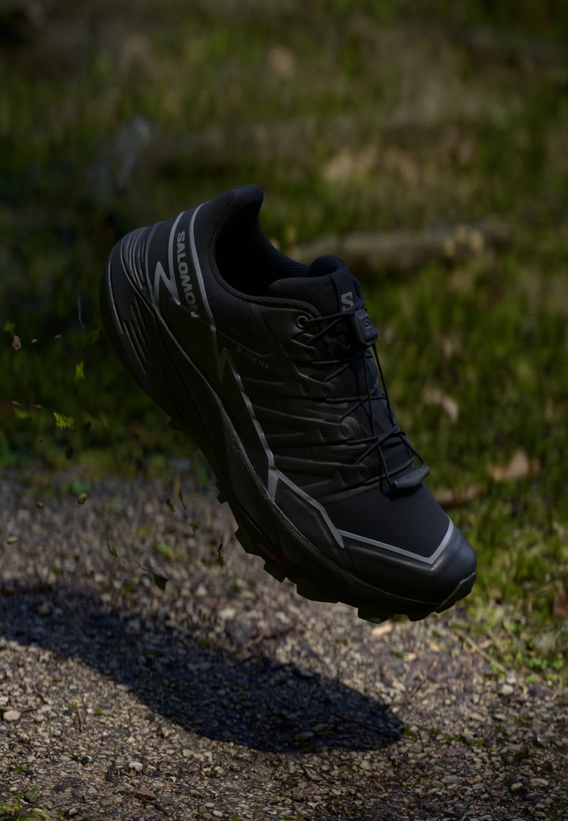 Zwarte Salomon trailrunningschoen die boven een rotsachtige ondergrond zweeft, met een achtergrond van groene bladeren. De zoolstructuur en het vetersysteem zijn zichtbaar.