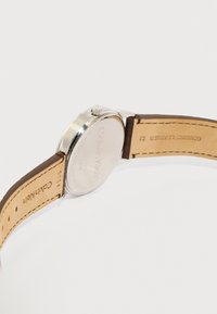 Ρολόι χειρός Calvin Klein με ασημένια θήκη, κορόνα και καφέ λουράκι από γνήσιο δέρμα, φωτογραφημένο από πίσω σε λευκό φόντο.