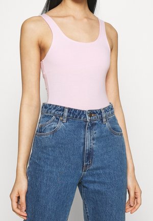 Femme portant un haut sans manches rose clair rentré dans un jean en denim bleu taille haute, debout avec les bras détendus le long du corps.