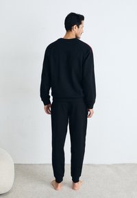 Schwarzes Sweatshirt und passende Jogginghose aus weichem Stoff. Das Sweatshirt hat einen roten Akzent entlang der Schulter. Schlichte, moderne Passform.
