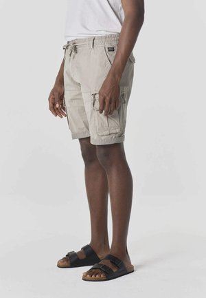 Hombre con pantalones cortos cargo beige con cintura de cordón y sandalias negras de doble correa, de pie sobre un fondo claro y liso.