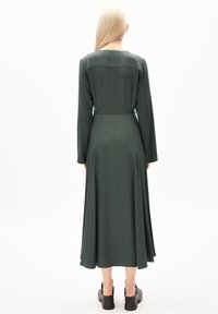 Pitkä, tummanvihreä mekko, jossa on pitkät hihat, vyö vyötäröllä, virtaava kangas ja suora leikkaus. Sileä pinta ja yksinkertainen muotoilu.