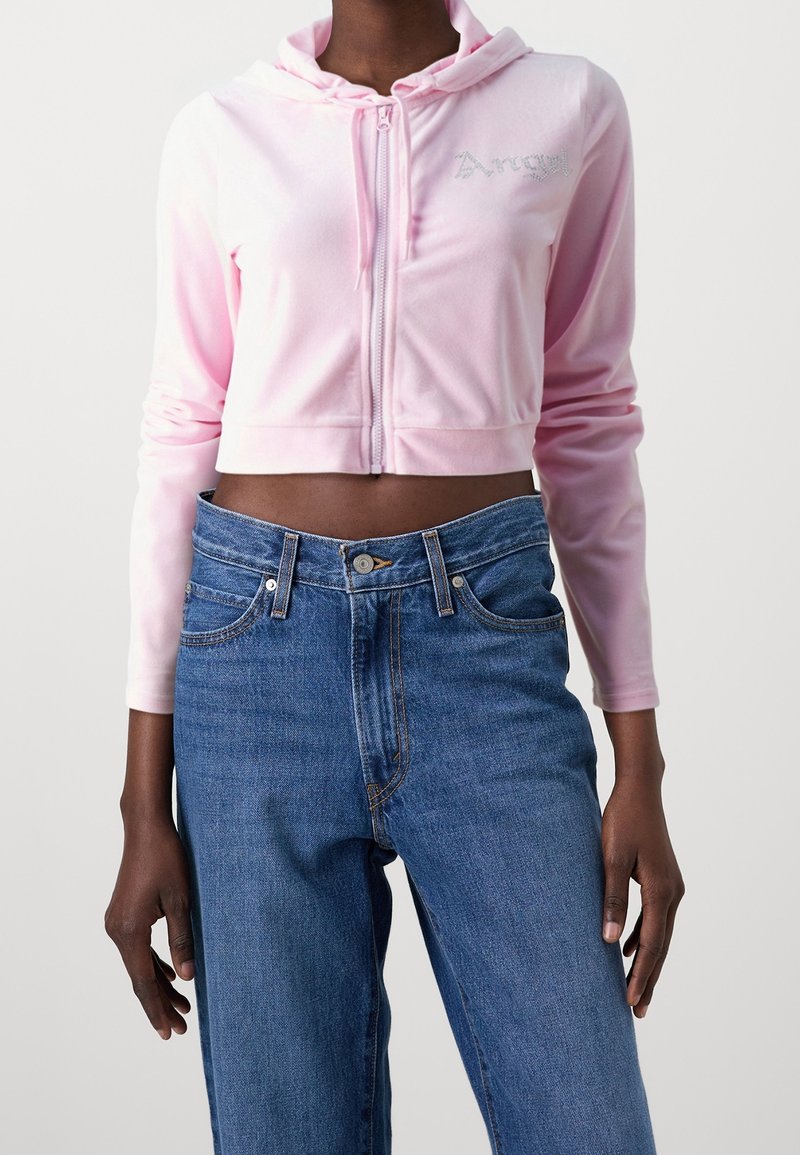 Personne portant un sweat à capuche court rose clair avec fermeture éclair et texte « Angel », ainsi qu'un jean taille haute bleu, sur un fond uni.