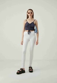Top cropped navy com detalhe em cordão, jeans skinny brancos e sandálias pretas chunky com tachas. Os acessórios incluem óculos de sol brancos e uma mala azul.