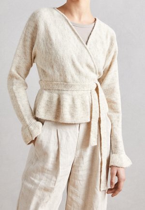 Persona che indossa un maglione beige con cintura sopra una canotta chiara e pantaloni larghi di lino chiaro su uno sfondo semplice.